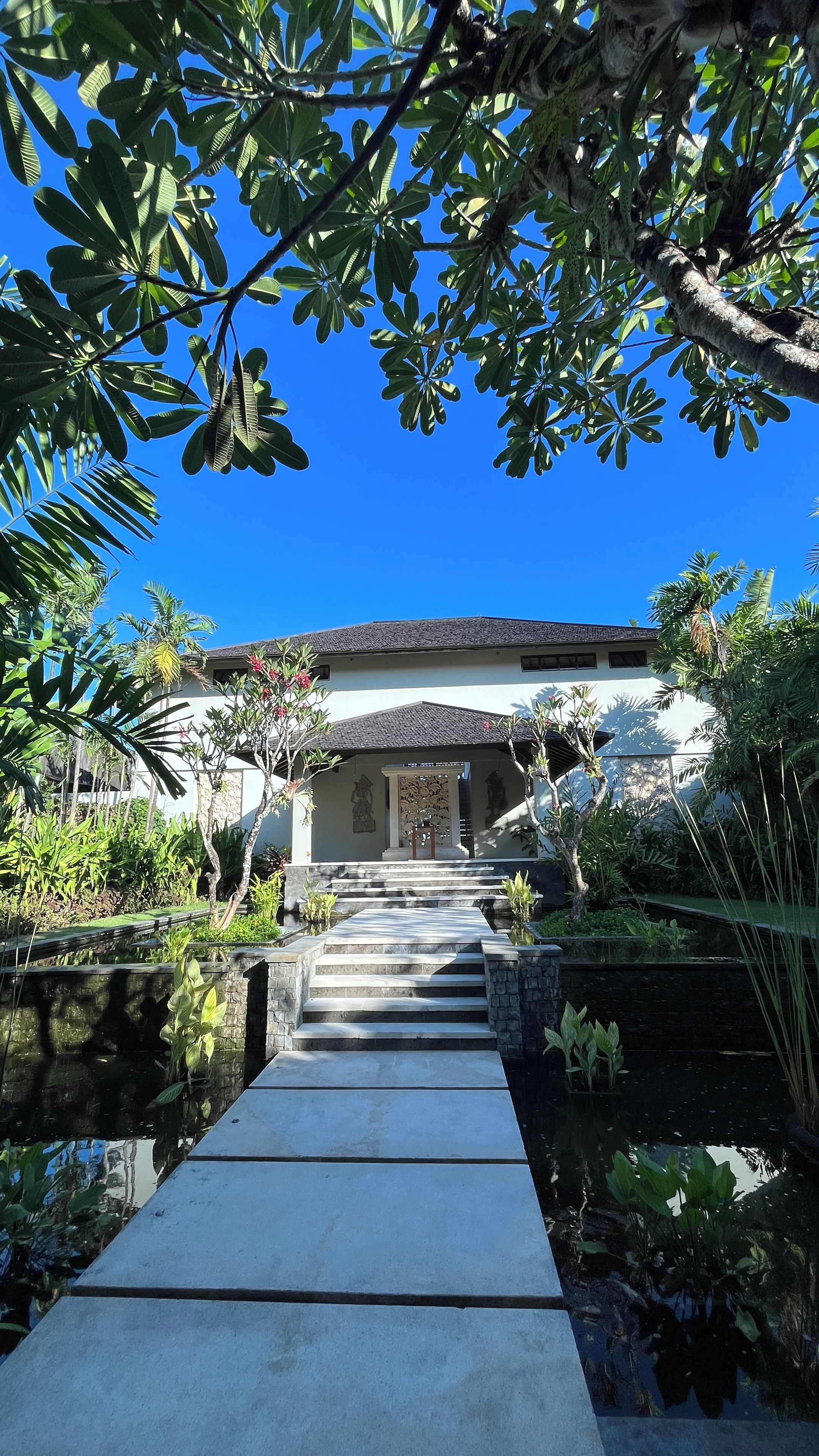 A pathway leading to a building with a tiled roof, surrounded by lush greenery and a small water feature.