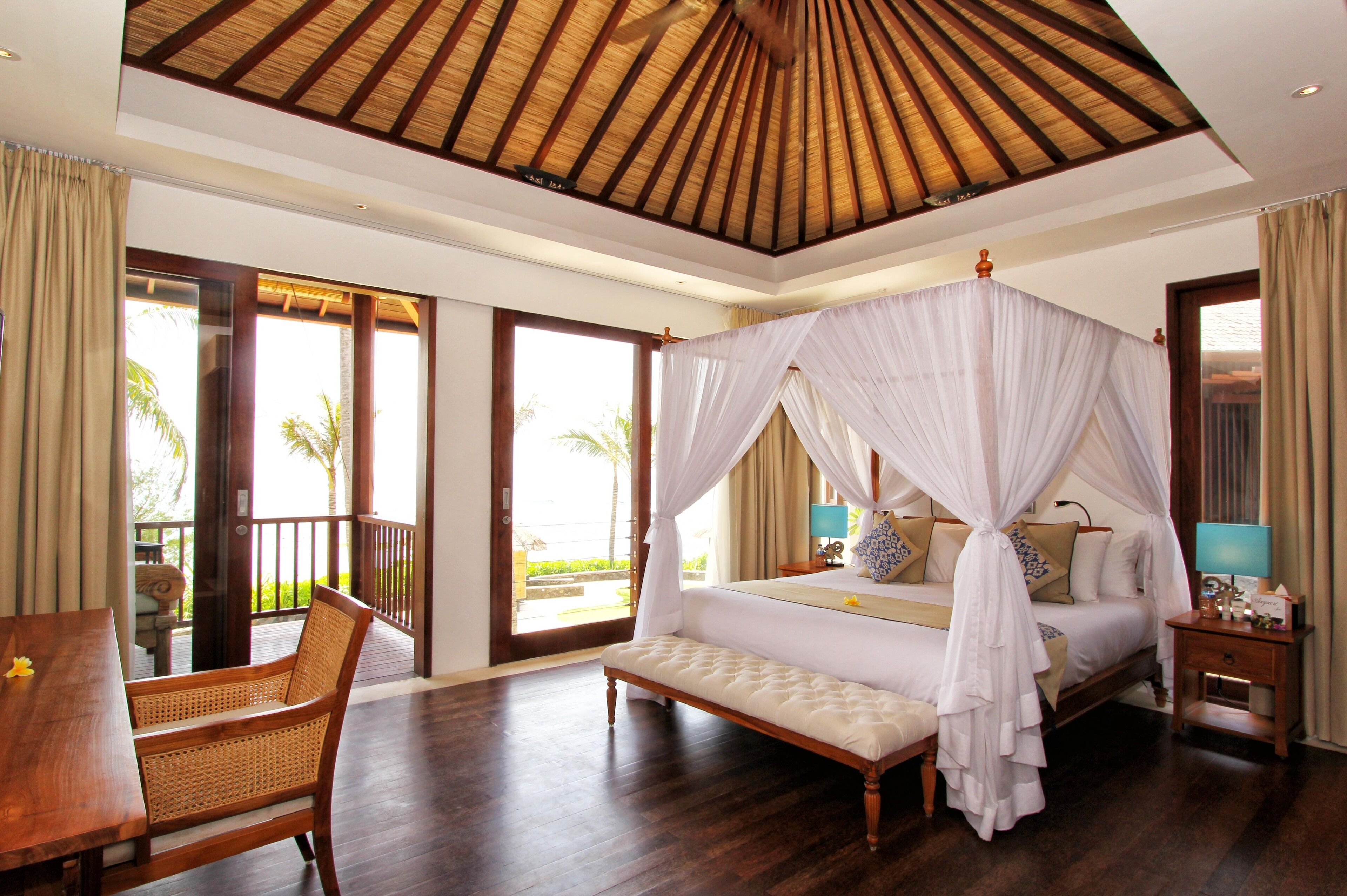 A spacious bedroom with a four-poster bed, a wooden bench, a bedside table, and a view of the outdoors.