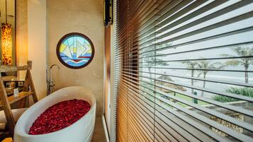 A bathroom with a bathtub filled with rose petals, a stained glass window, and a view of palm trees through window blinds.