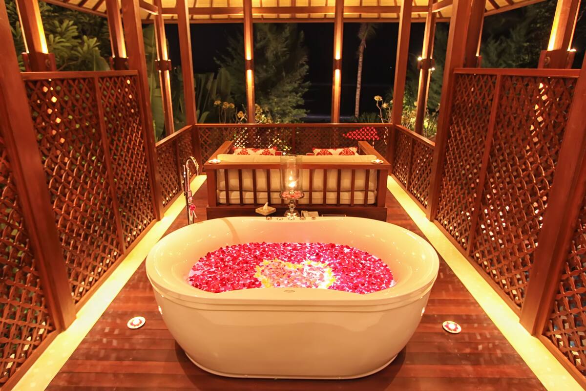 Ein Wellnessbereich mit einer großen Badewanne, gefüllt mit Rosenblättern, Holzgitterverkleidungen und einer Sitzecke mit Kissen.