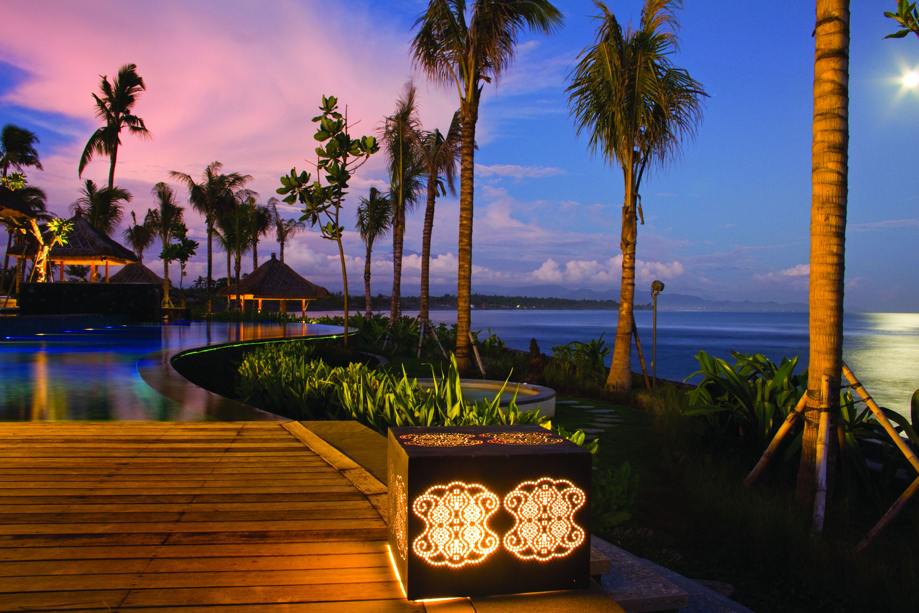 Une terrasse en bois agrémentée d’un luminaire décoratif, de palmiers et offrant une vue sur l’océan.