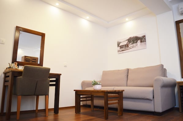 Una sala de estar con un sofá, una mesa de madera, una silla, una mesita con una planta y un cuadro en la pared.