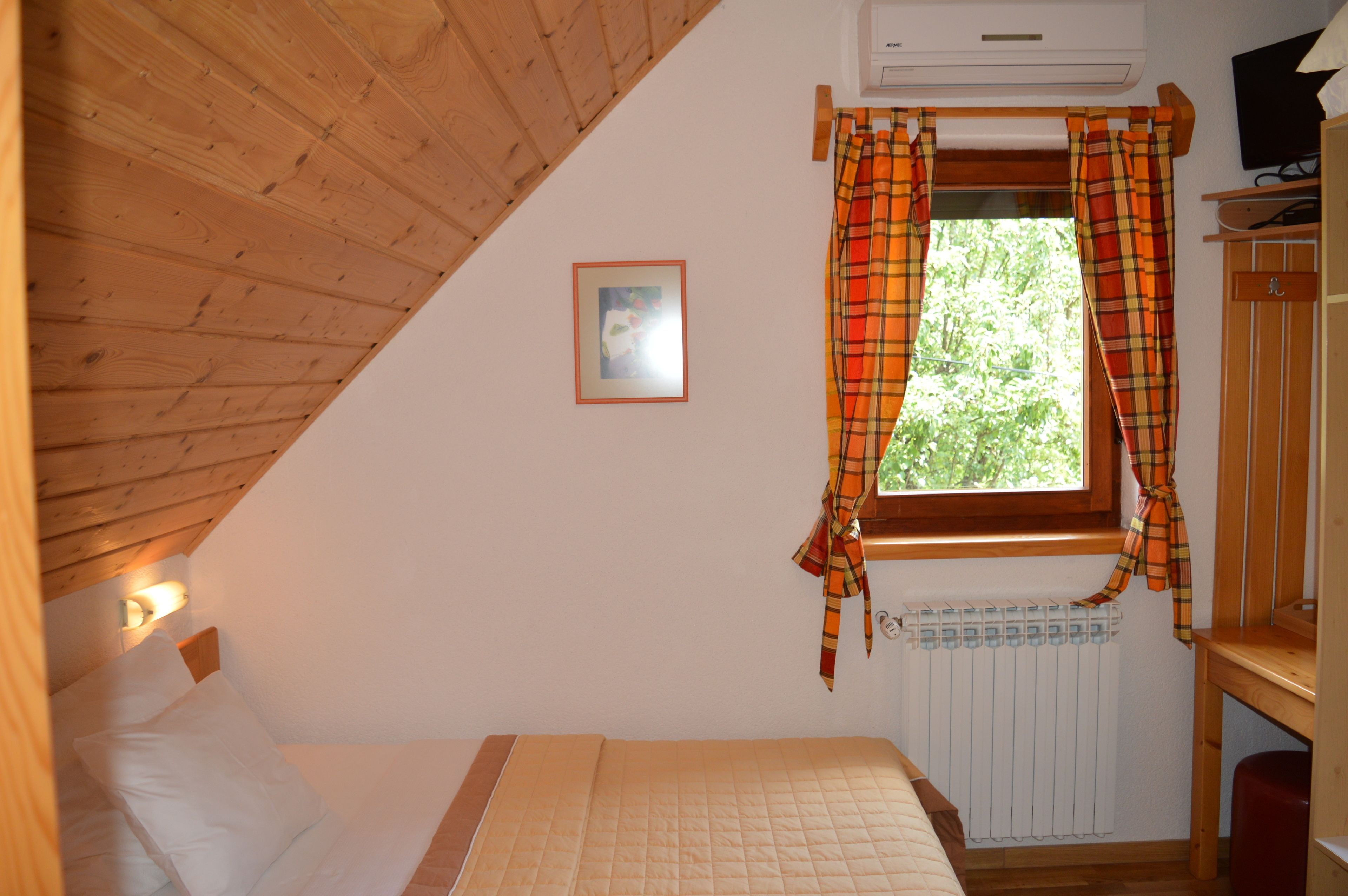 A bedroom with a wooden ceiling, a bed with white bedding, a window with plaid curtains, a radiator, and a small picture on the wall.