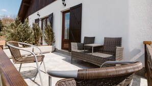 An outdoor seating area with wicker furniture and a wooden table, a white building with wooden shutters, and a potted plant.