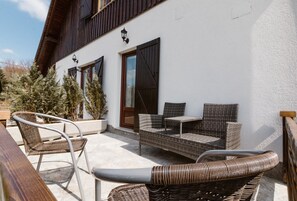 An outdoor seating area with wicker furniture and a wooden table, a white building with wooden shutters, and a potted plant.