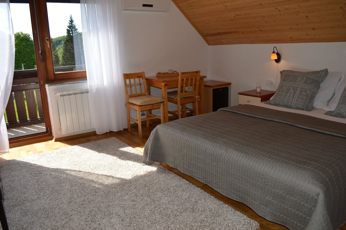 A bedroom with a bed, a desk, and a chair. There is a window with curtains, a radiator, and a nightstand with a lamp.