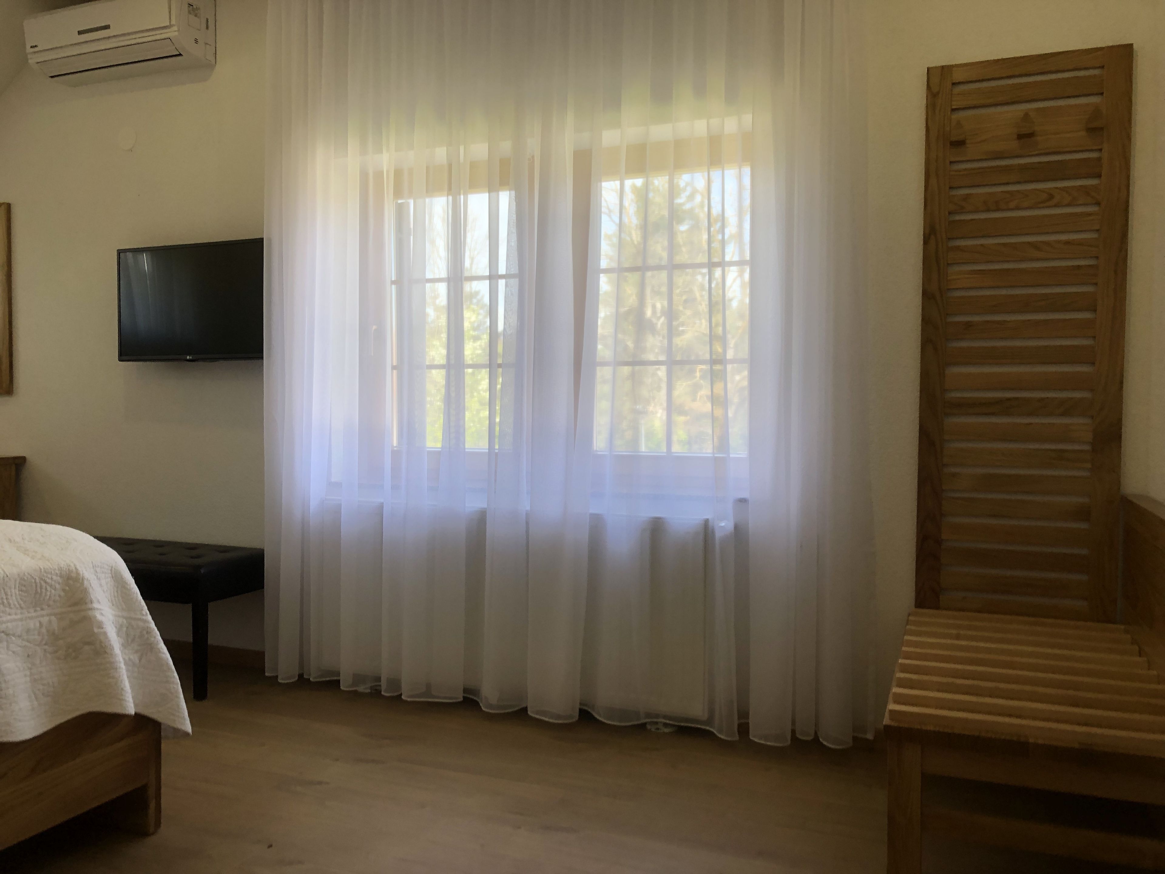 A room with a bed, a television, two windows with sheer curtains, a wooden bench, and a wooden staircase.