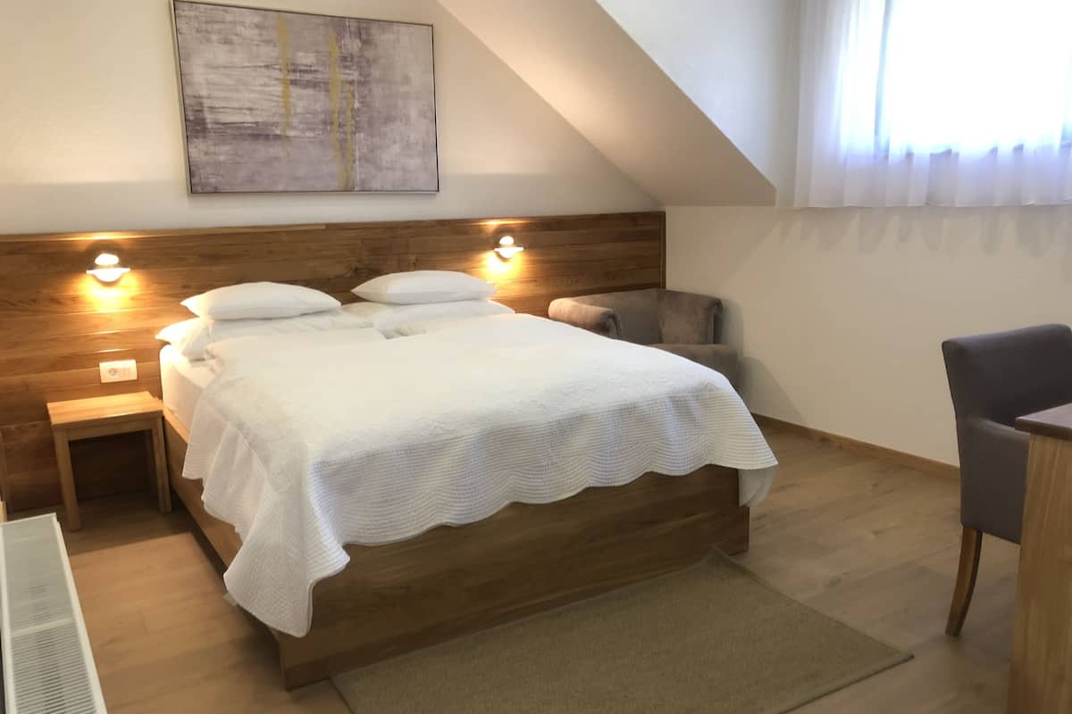 A bedroom with a wooden headboard, a bed with white linens, two bedside lamps, a small nightstand, a chair, and a large abstract painting on the wall.