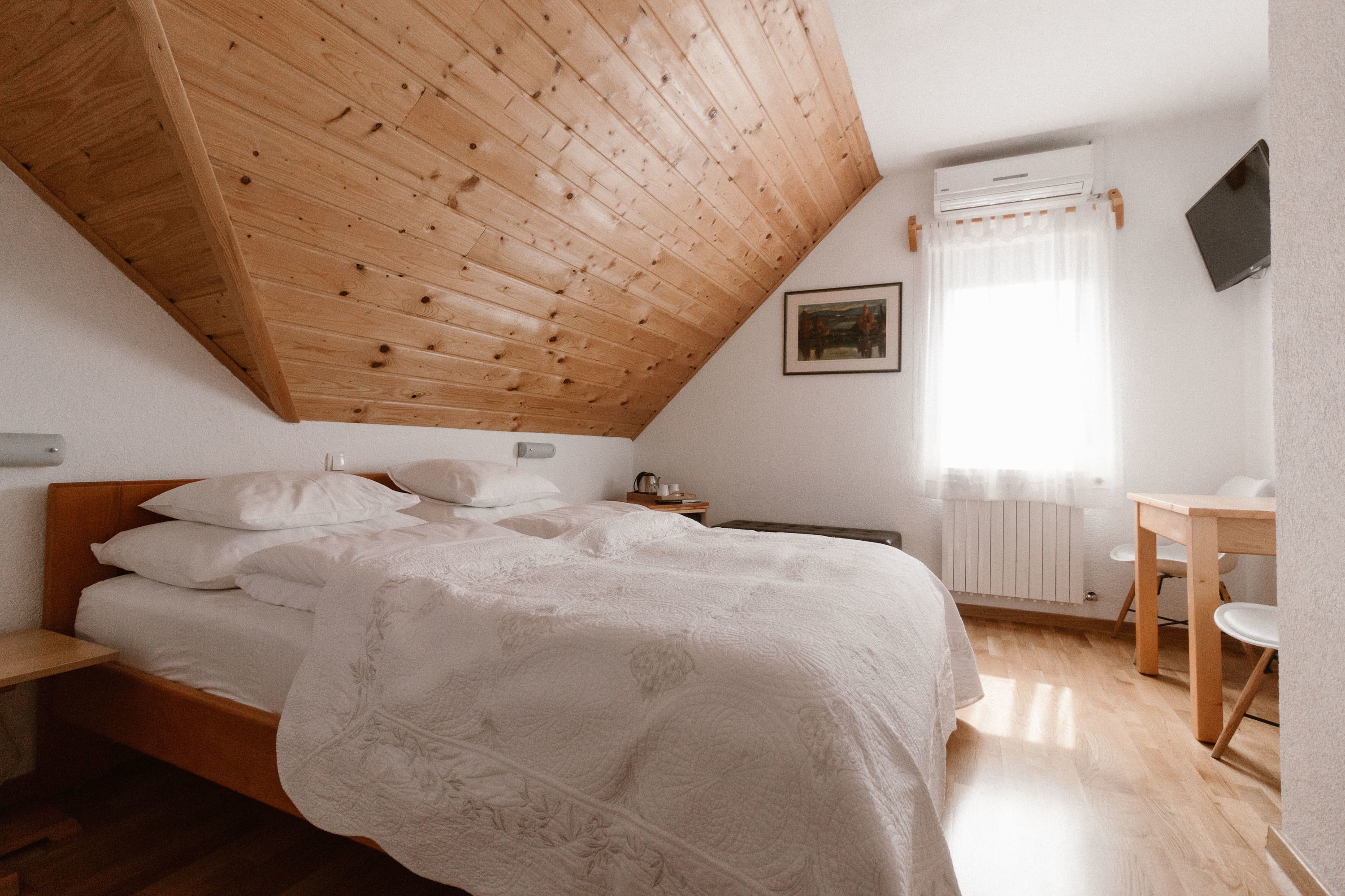 A bedroom with a sloped ceiling, a bed with white linens, a bedside table, a small table with chairs, a radiator, and a TV mounted on the wall.