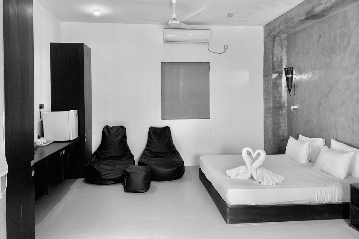 A modern hotel room with a large bed, a ceiling fan, an air conditioning unit, a small desk with a chair, and two bean bags.