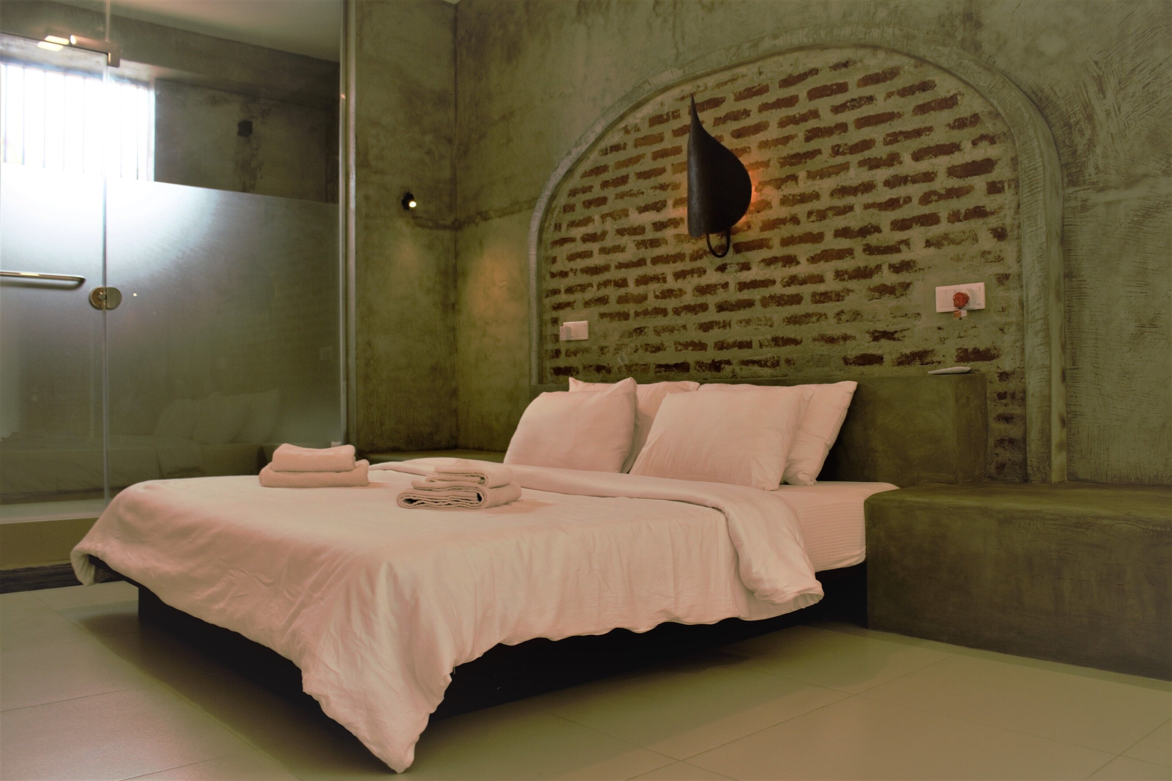 A bedroom with a bed, a bench, a glass shower, and a brick-patterned wall.