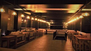 A dimly lit restaurant with long tables and benches, overhead lighting, and a view of the cityscape in the background. - Handagedara Resort (Mirissa)