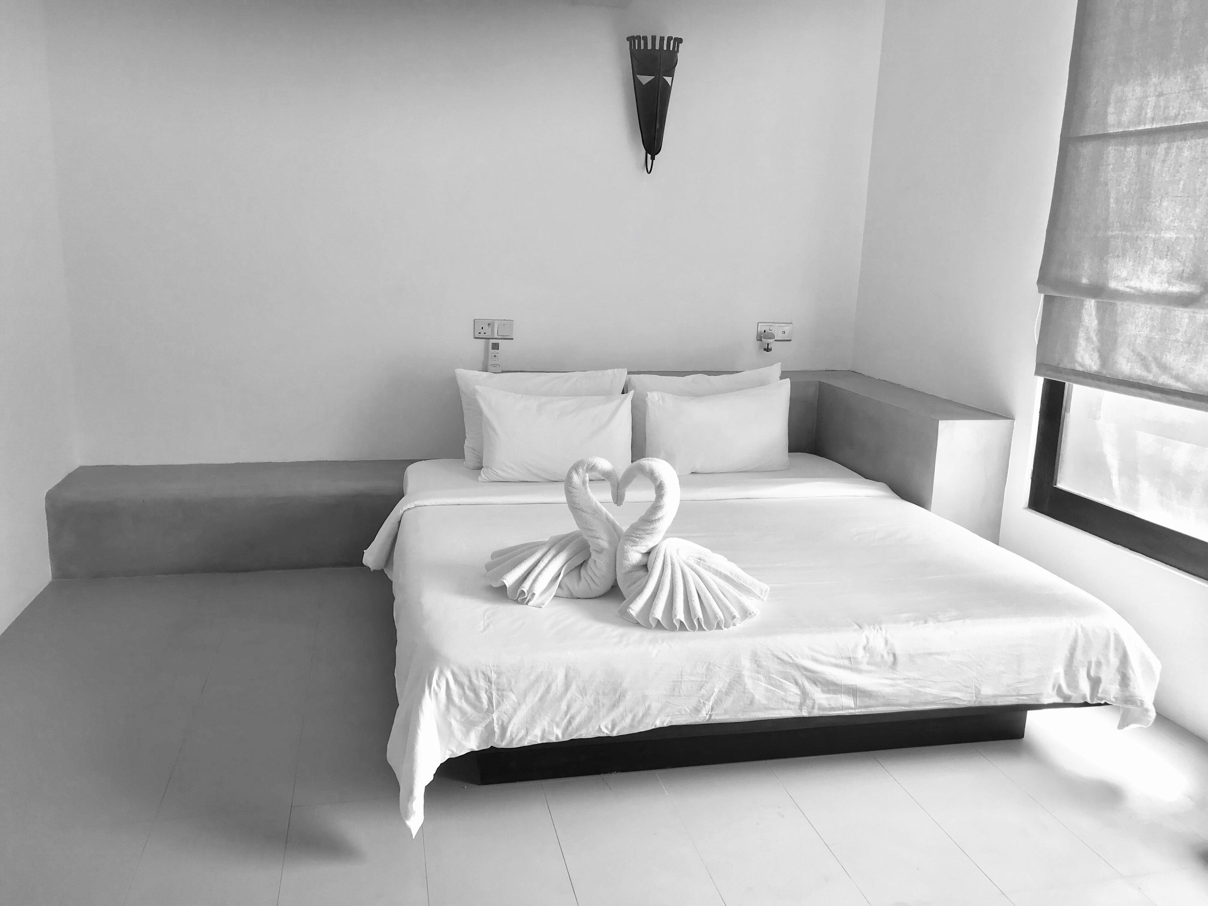 A modern hotel room with a large bed, two bedside tables, a wall-mounted light fixture, and a window with a blind.