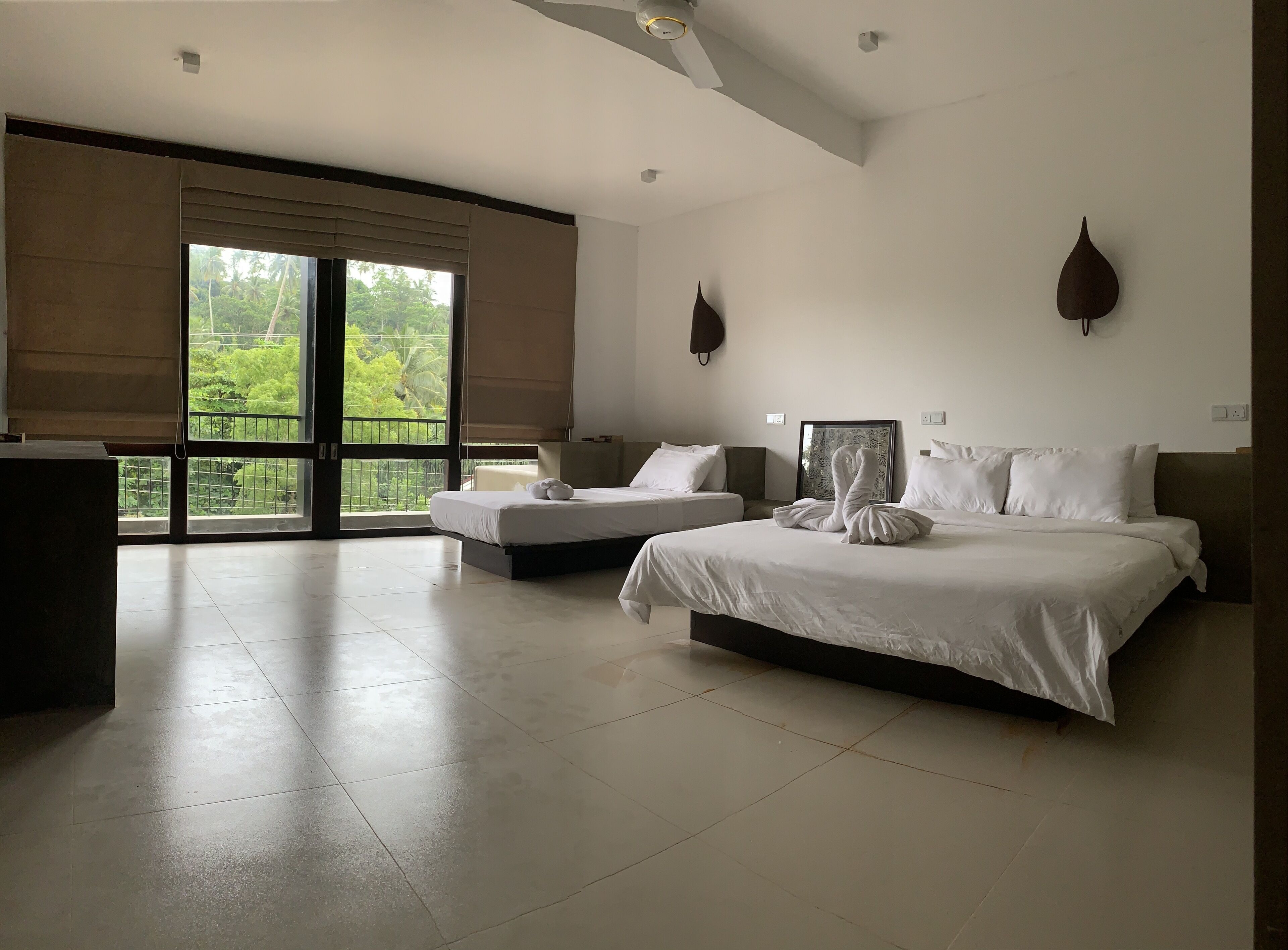 A spacious hotel room with a large bed, a smaller bed, and a sofa. There is a balcony with a view of greenery.
