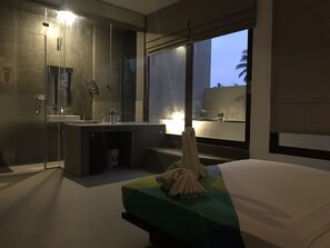 Una habitación de hotel moderna con cabina de ducha de vidrio, una mesita de noche con lámpara y un amplio ventanal con vistas a palmeras.