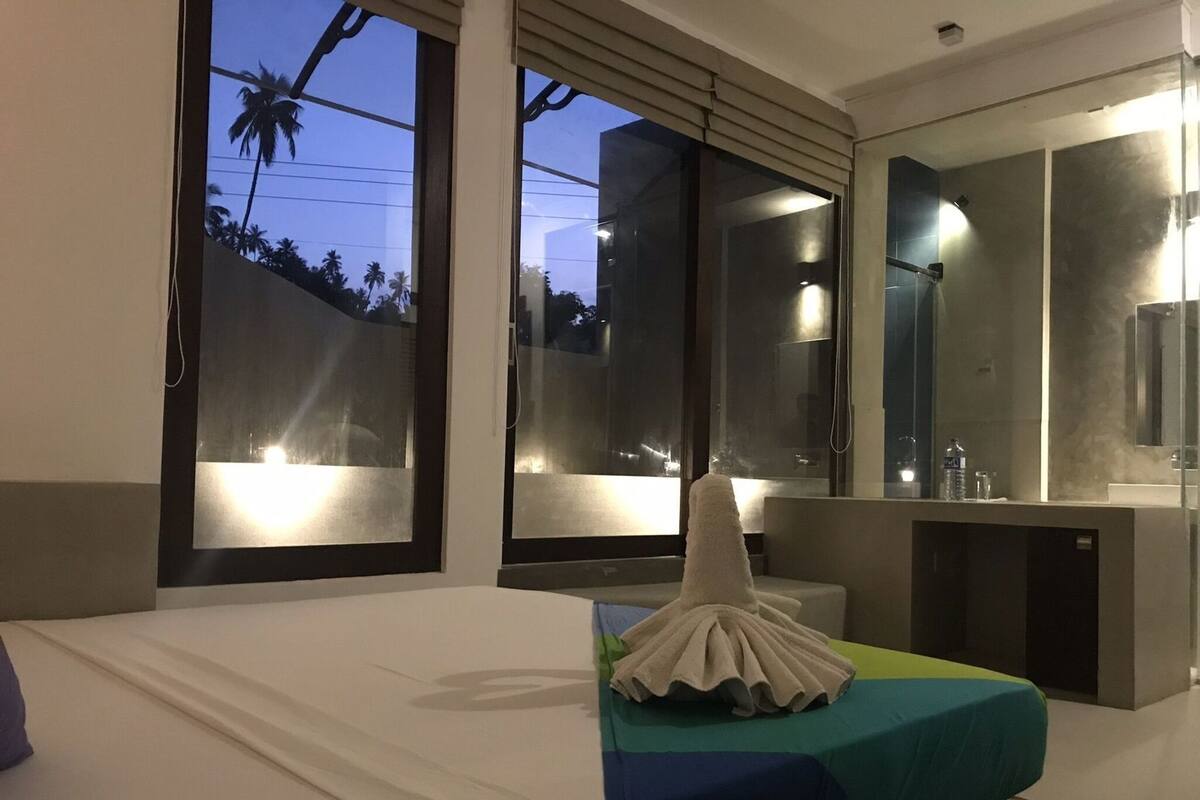 A modern hotel room with a large bed, a bathroom with a mirror and sink, and large windows with a view of palm trees.