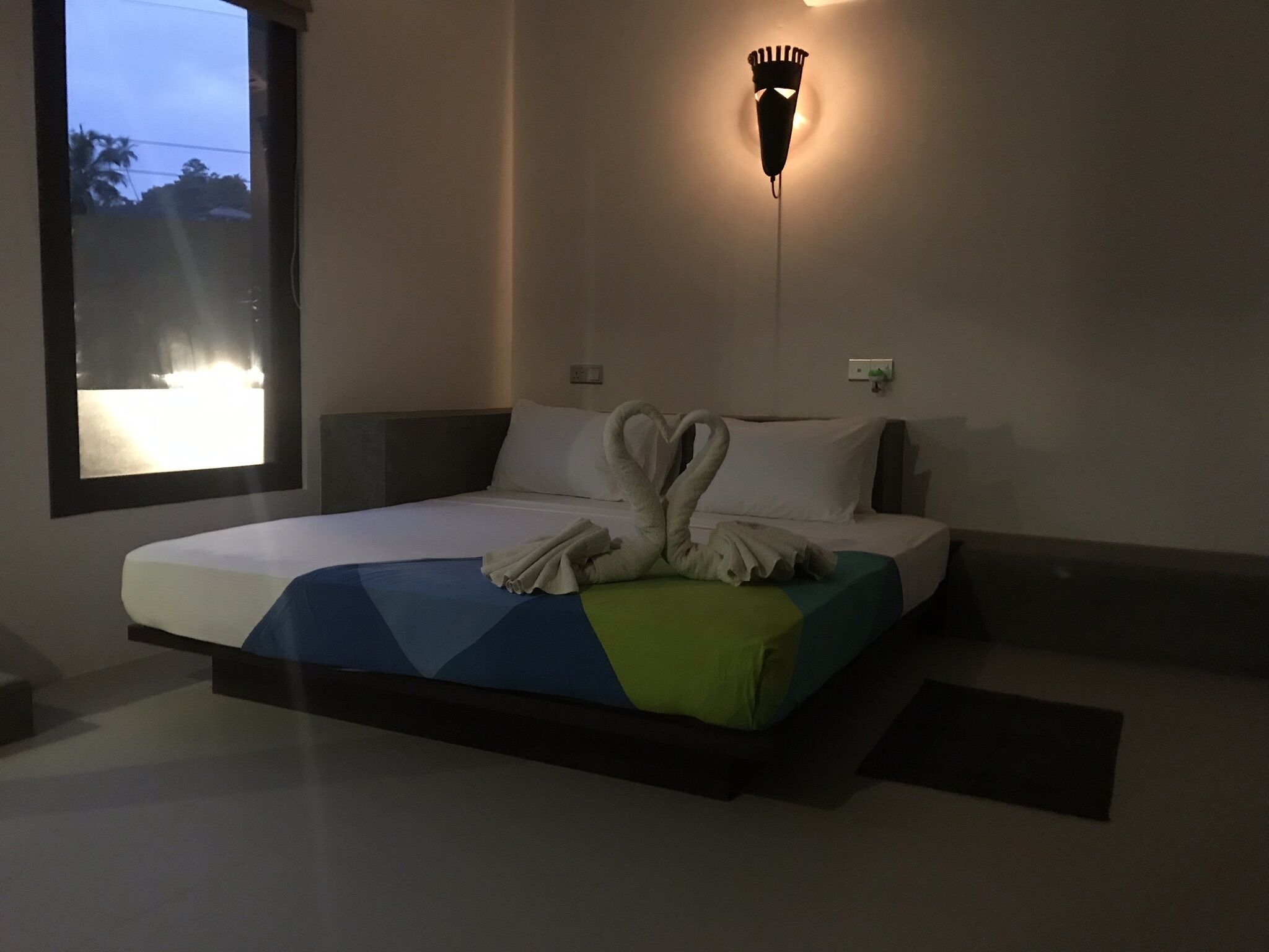 A hotel room with a bed, a wall-mounted light fixture, and a window with a view of palm trees.