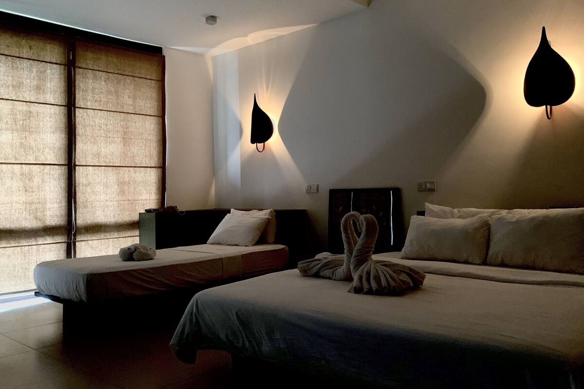 A modern hotel room with a large bed, a smaller bed, and a seating area. There are wall-mounted lights and a large window with blinds.