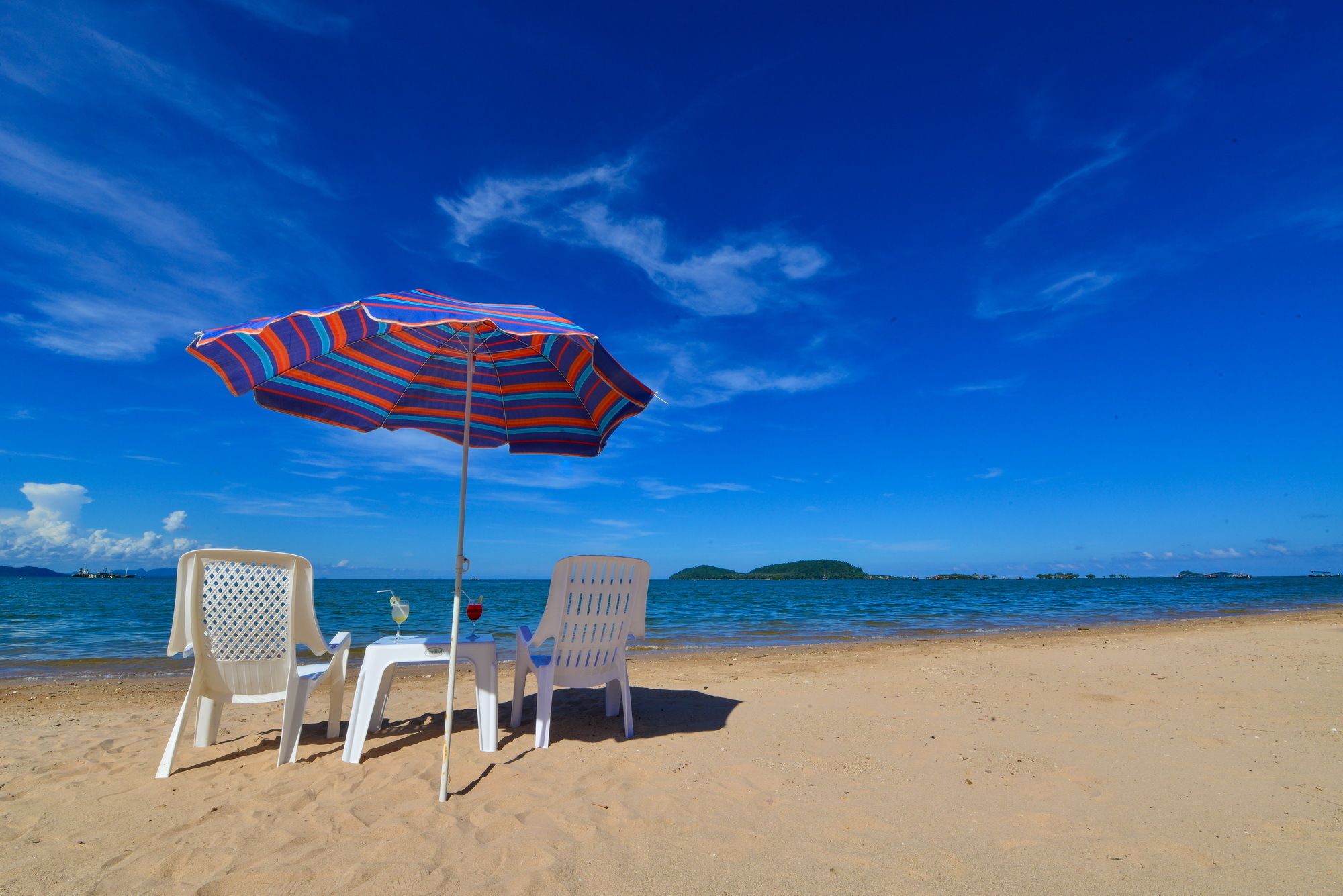 A beach with two white plastic chairs and a striped umbrella.