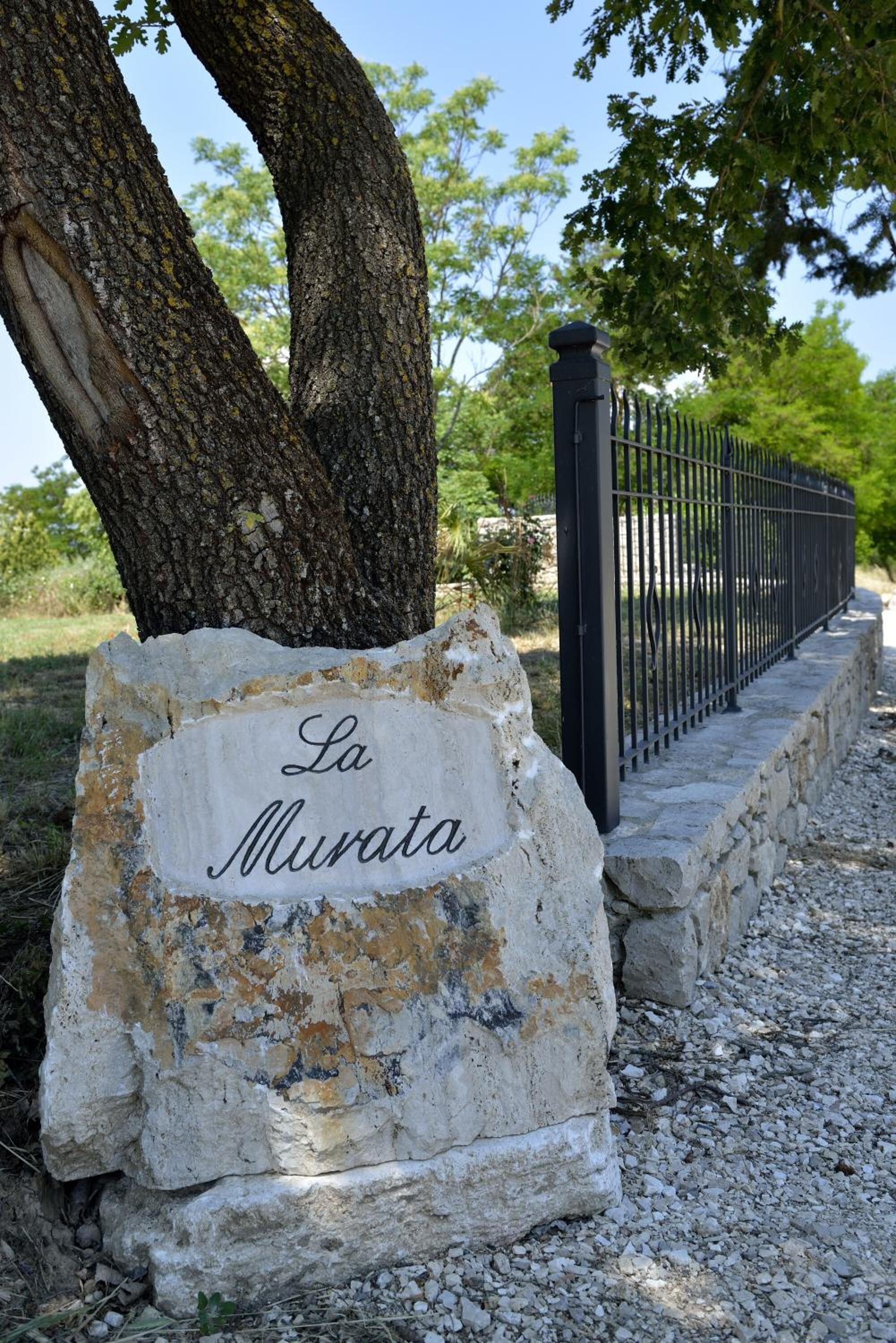 Un letrero de piedra con la inscripción 'La Murata', ubicado al lado de un sendero de grava y una valla metálica.