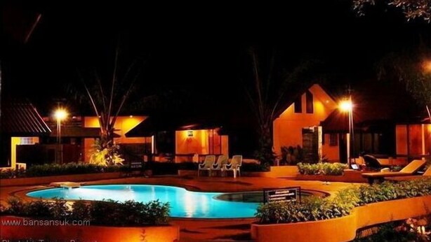Vista nocturna de un resort con piscina, sillas de descanso y edificios bien iluminados.