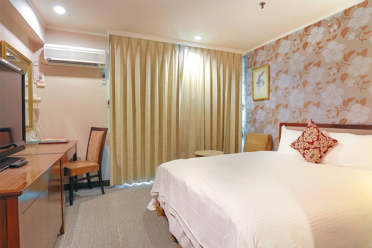A hotel room with a bed, a desk, a chair, an air conditioning unit, and a curtain.