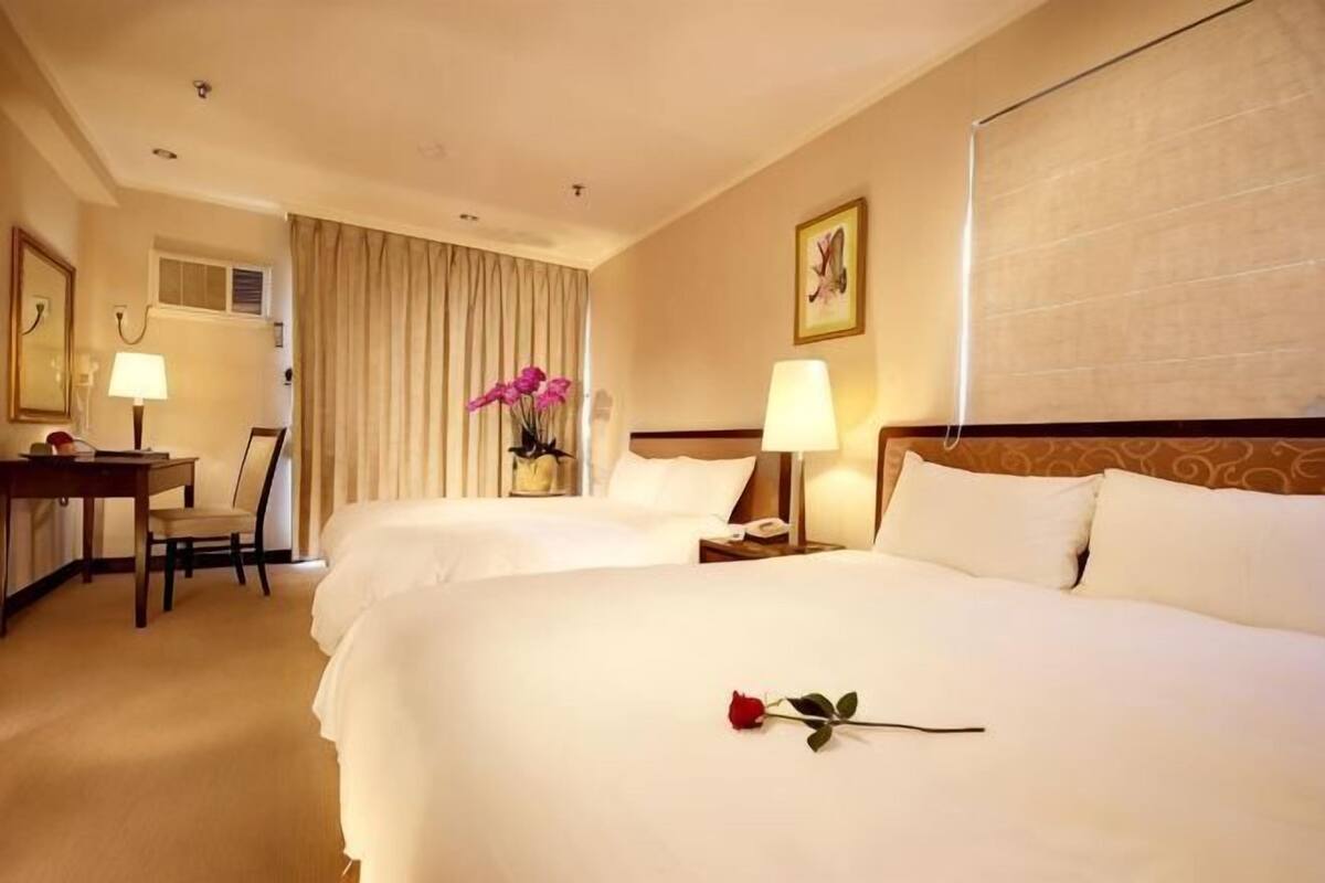 A hotel room with two beds, a desk, a chair, a lamp, and a vase of flowers.