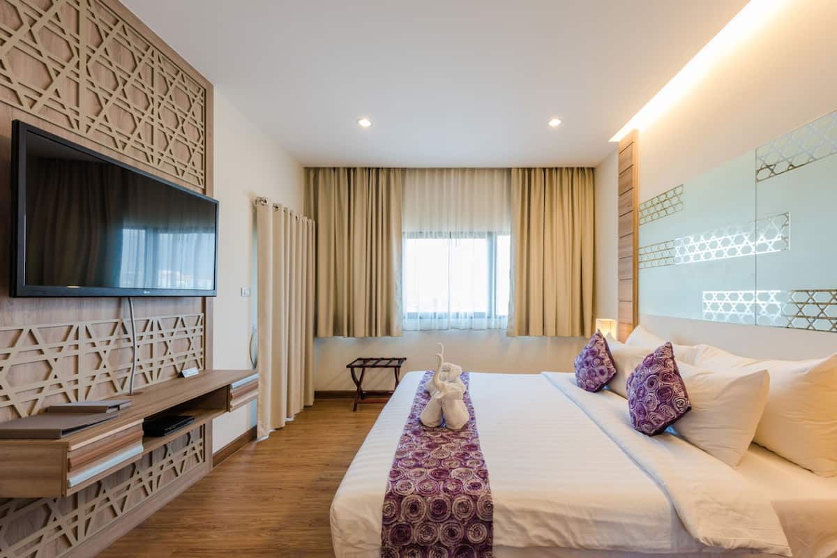 Ein Hotelzimmer mit einem großen Bett, einem an einer Holzvertäfelung montierten Flachbildfernseher, einem kleinen Tisch und einem Fenster mit Vorhängen.