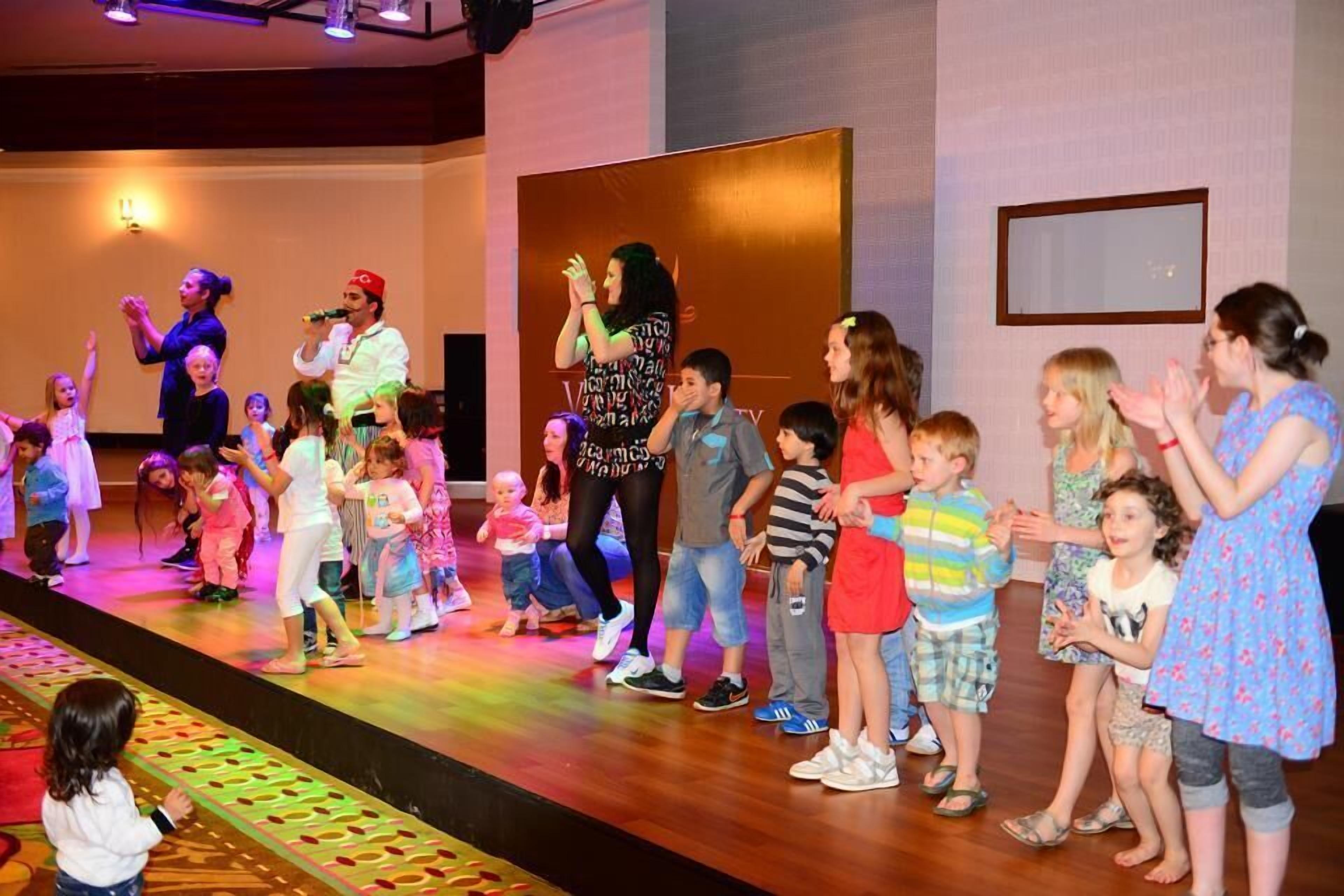 A stage with children and adults, some holding microphones, in a room with a wooden floor and a large screen in the background.