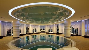 A modern indoor swimming pool with a central circular seating area, surrounded by large windows and curtains. - Vikingen Infinity Resort & Spa - All Inclusive (Alanya)
