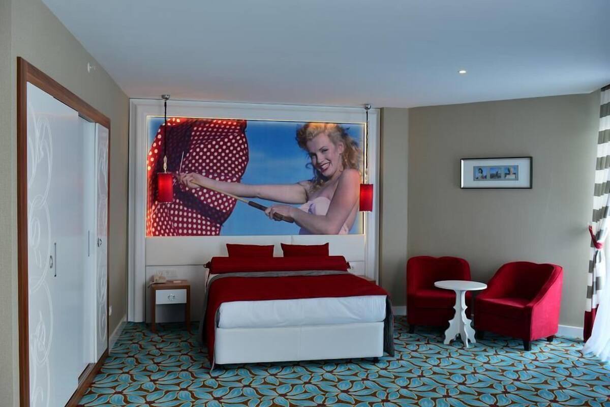 A hotel room with a bed, two red chairs, a small table, and a large window with a picture.