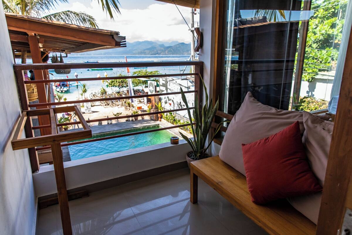 A balcony with a view of a pool and beach, a wooden bench, and a red pillow.