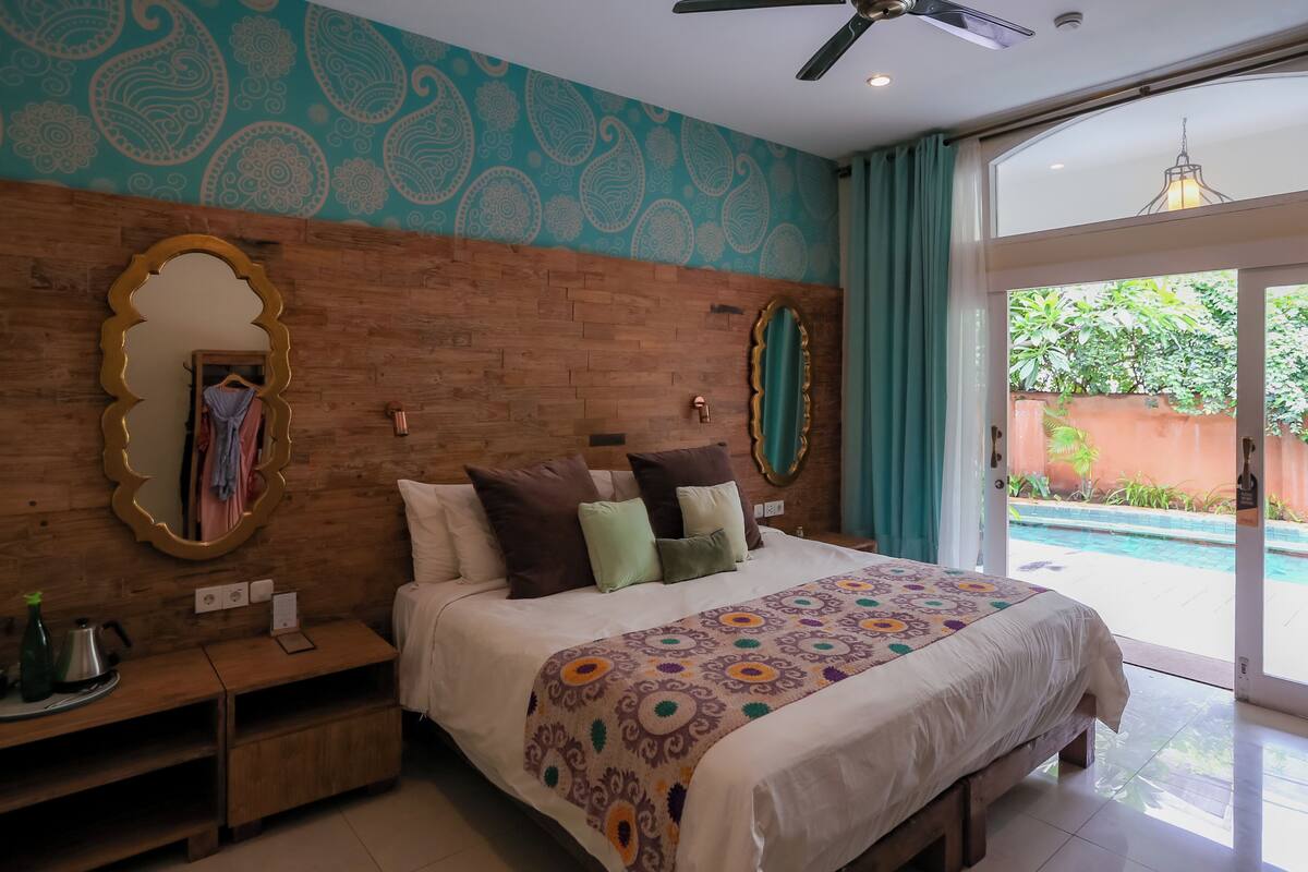 A bedroom with a wooden headboard, a large mirror, a bed with multiple pillows, a bedside table with a bottle and a kettle, and a view of a swimming pool through a glass door.