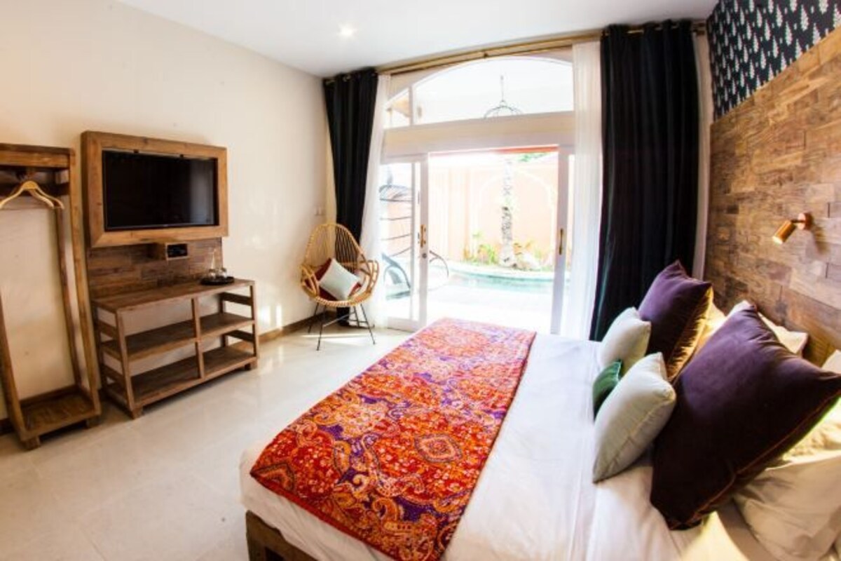 A bedroom with a large bed, a TV mounted on a wooden shelf, a chair, and a view of the outdoors.