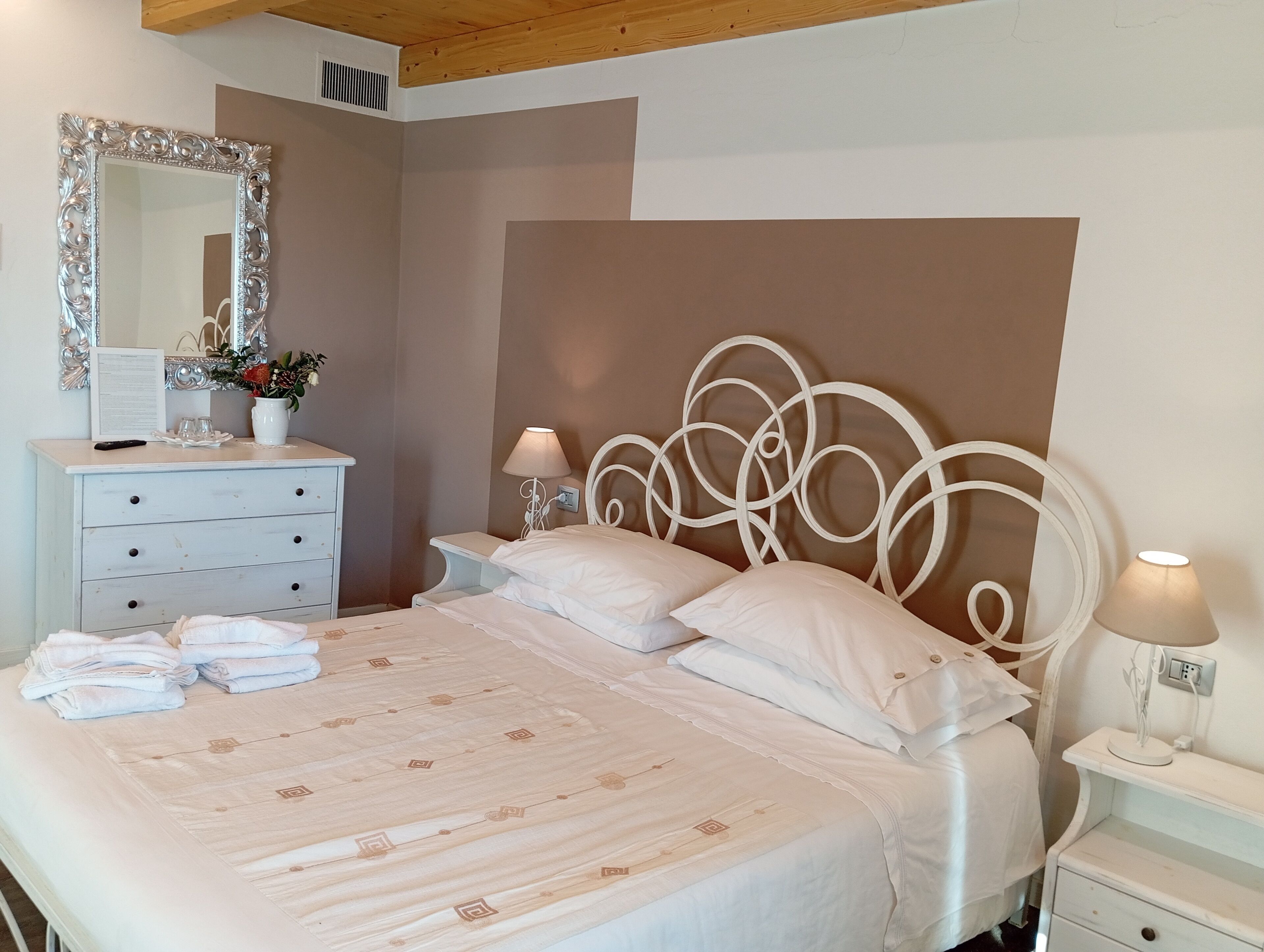 Un lit bien fait, recouvert d’une couvre-lit à motifs, une commode blanche avec des tiroirs, un miroir et deux lampes de chevet.