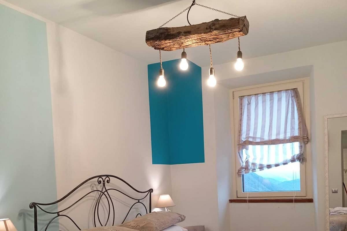 A bedroom with a bed, bedside lamps, a hanging light fixture, a window with curtains, and a view of the outdoors.
