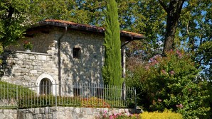 A stone building with a tiled roof, a metal fence, and lush greenery including a tall tree and flowering bushes.