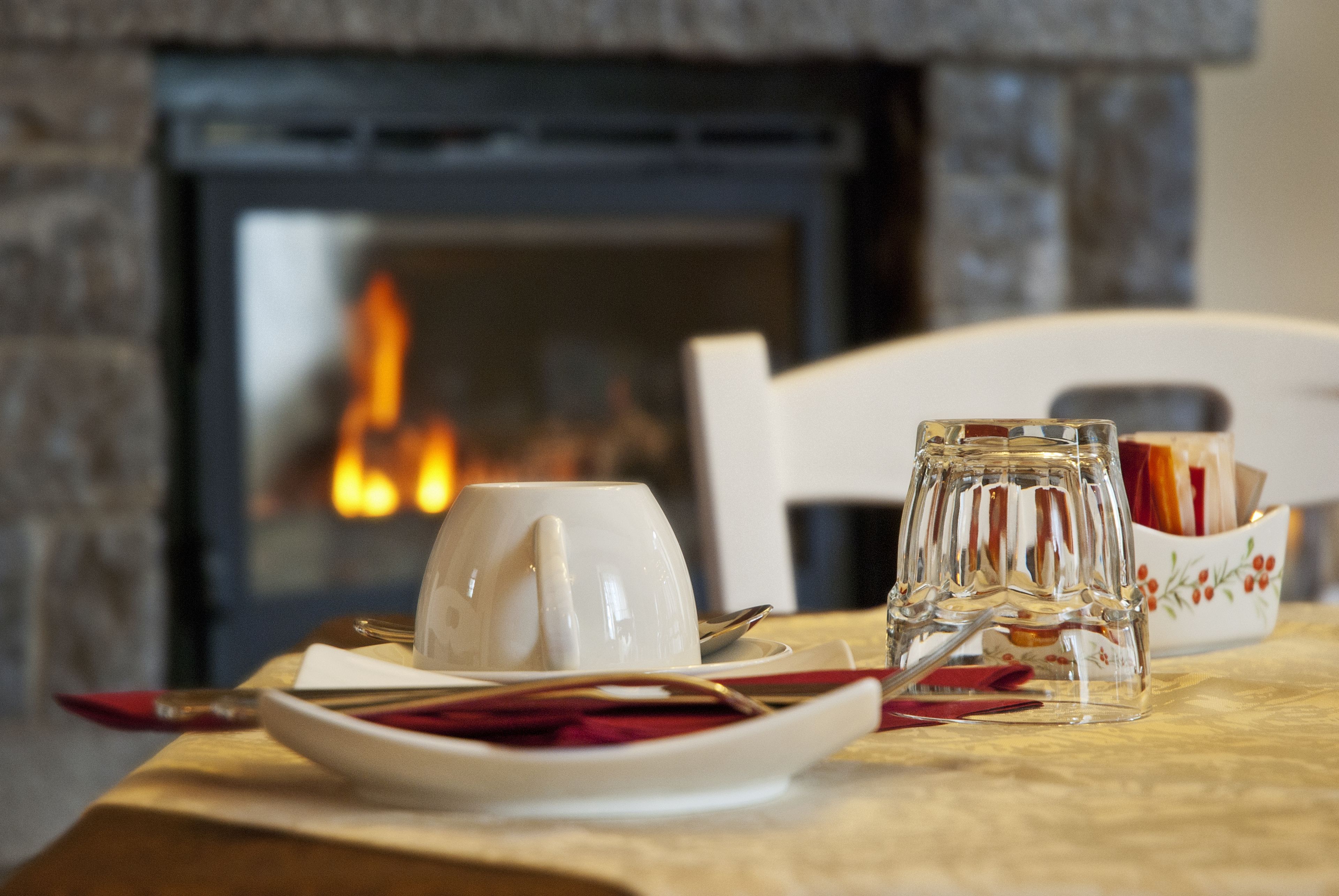 A dining table with a white cup, saucer, and glass, set with cutlery and napkins, in front of a fireplace.