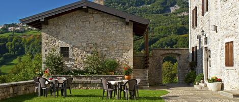 Un bâtiment en pierre doté d’un espace repas et de sièges en plein air, entouré de verdure et avec une chaîne de montagnes en toile de fond.