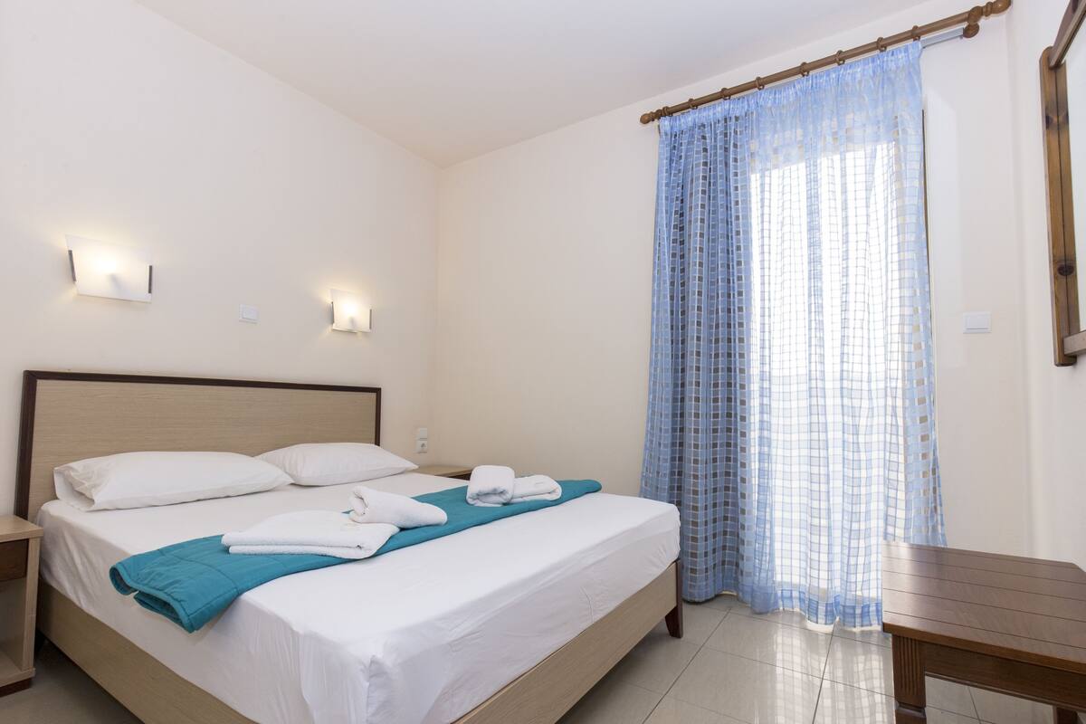 A neatly made bed with white linens and a teal throw, a wooden nightstand, a small table, and a window with a blue checkered curtain.