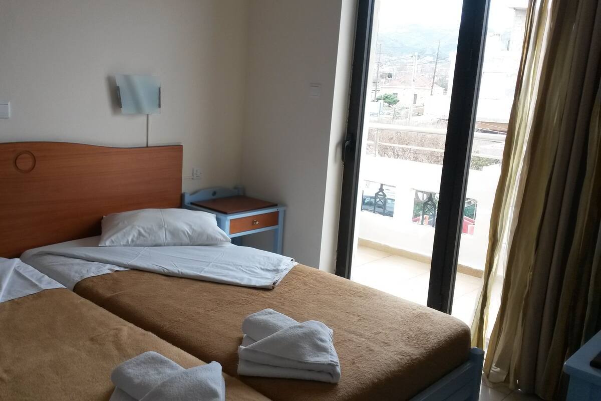 A hotel room with two beds, a nightstand, a lamp, and a view of the city through the window.