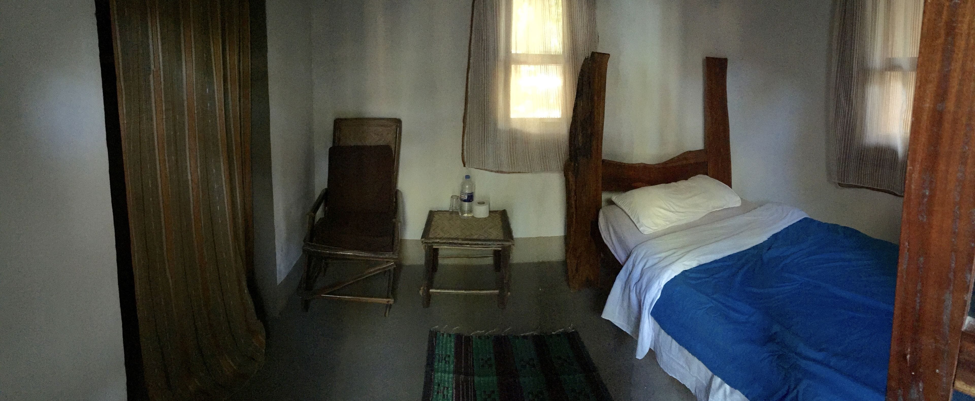Een kleine, zwak verlichte kamer met een enkel bed, een stoel, een tafeltje met een fles en een pak zakdoekjes, en een raam met gordijnen.