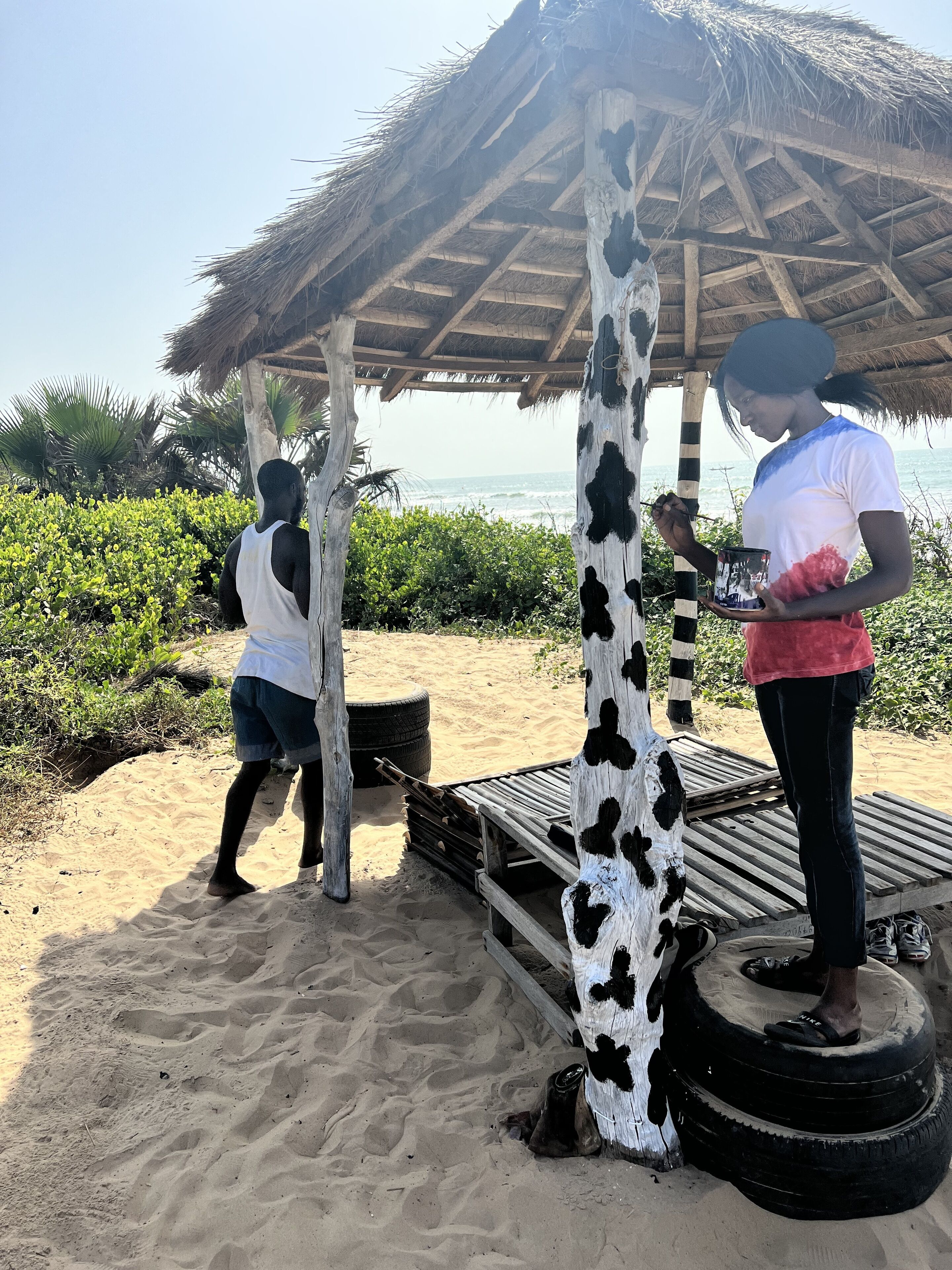A beach scene with a thatched-roof structure, a tire bench, and a person painting a wooden post.