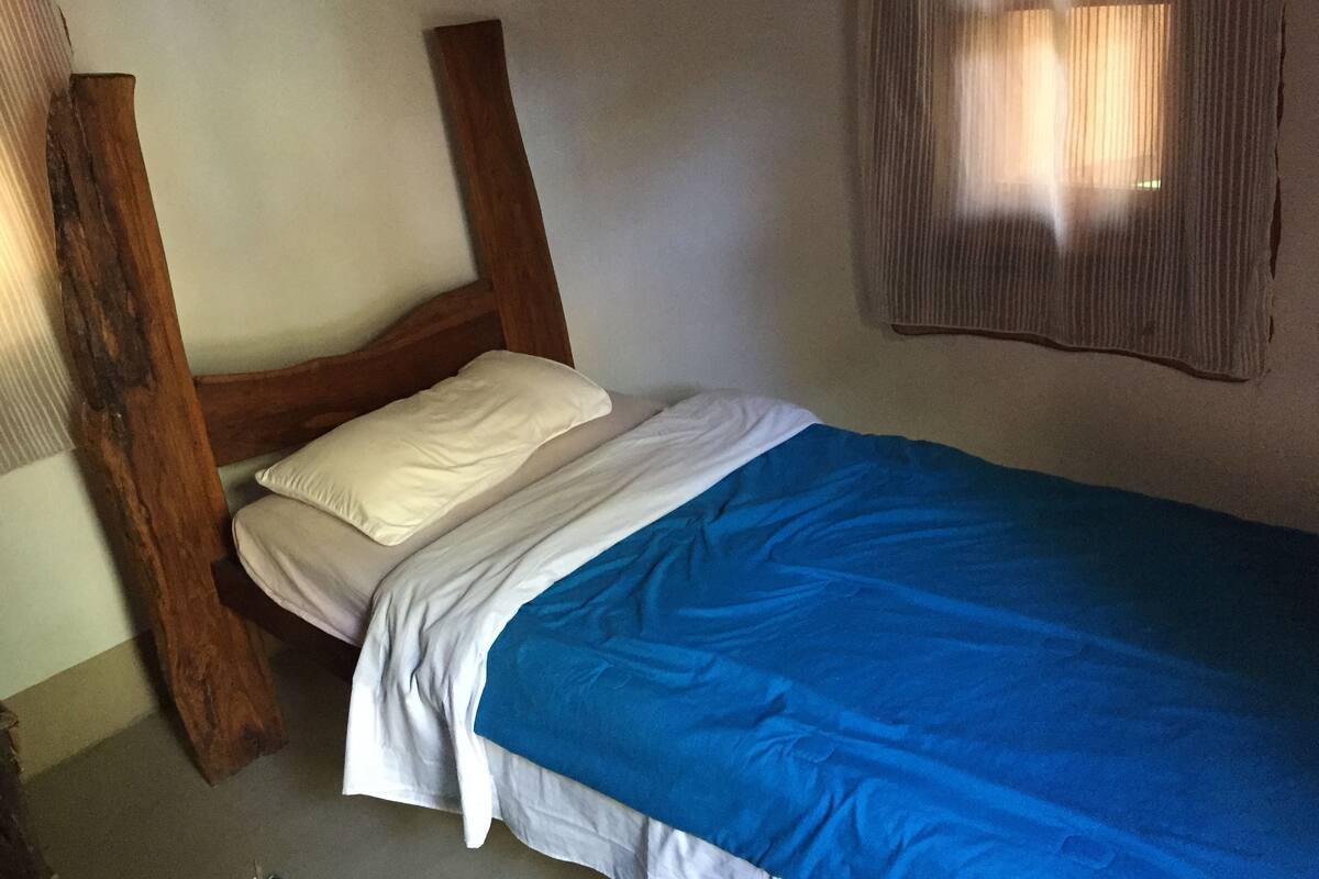A single bed with a wooden headboard, white and blue bedding, and a window with blinds.