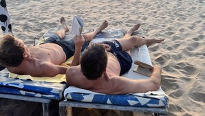 Dos personas descansando en sillas de playa bajo una sombrilla, con un toro y olas del océano al fondo.