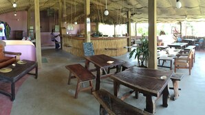 Un restaurante rústico con mesas y bancos de madera, una zona de bar y un techo de paja.