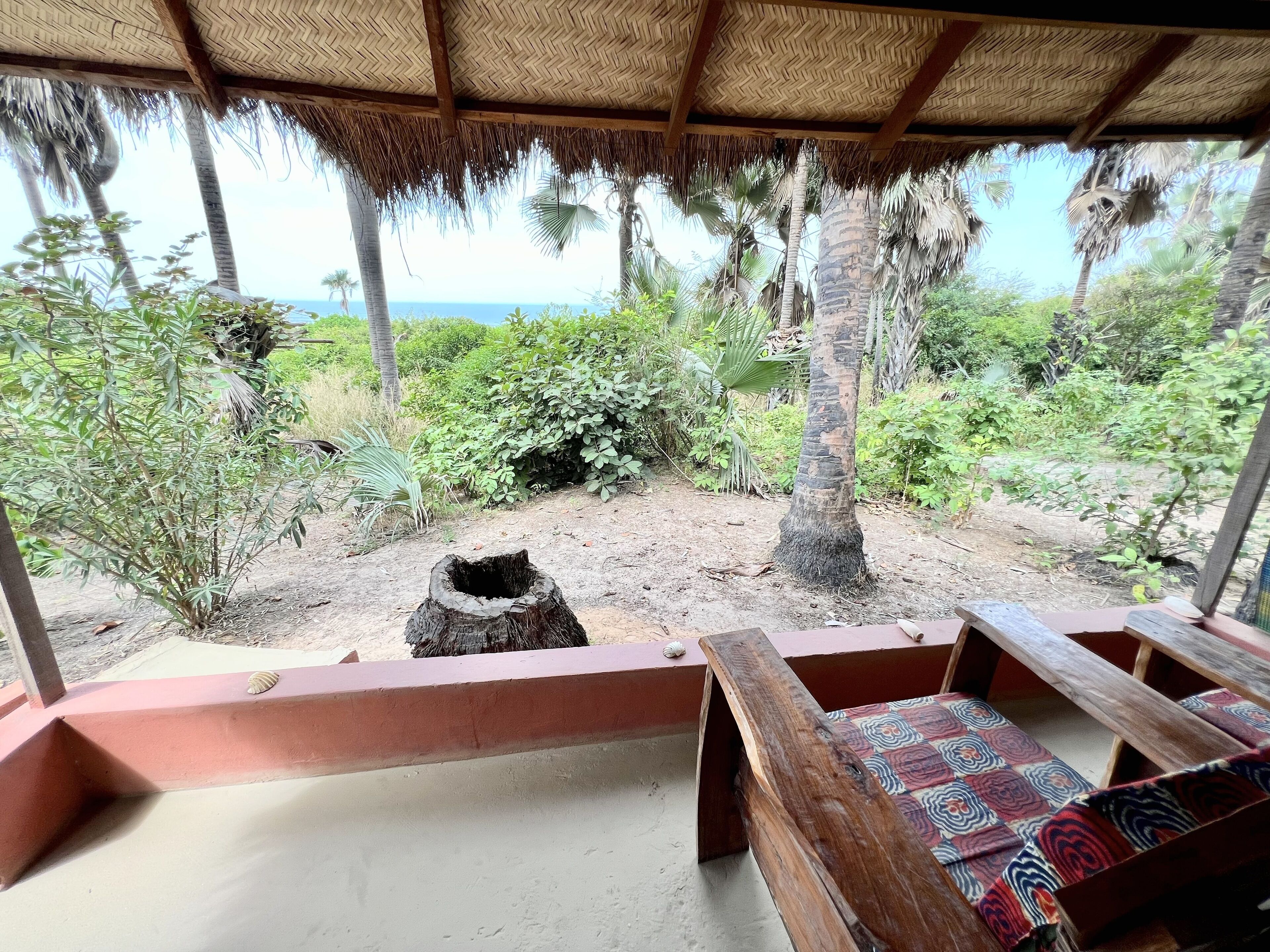 A thatched-roof outdoor seating area with benches, surrounded by lush greenery and a view of the ocean.