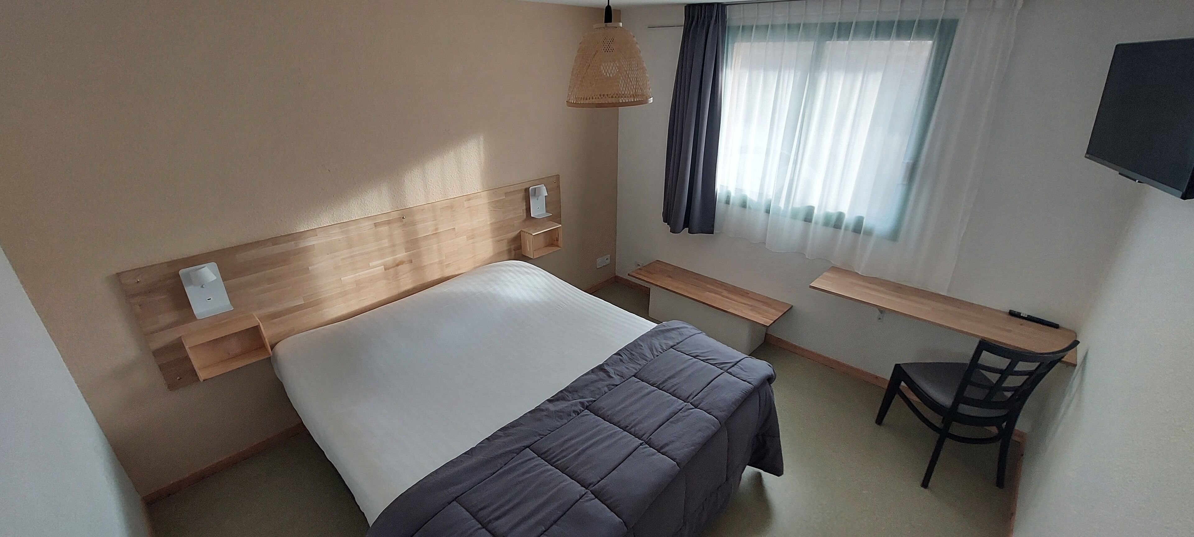 A hotel room with a wooden headboard, a bed with a grey duvet, a bedside table, a desk, a chair, and a TV mounted on the wall.