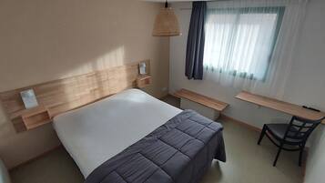 A hotel room with a wooden headboard, a bed with a grey duvet, a bedside table, a desk, a chair, and a TV mounted on the wall.