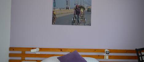 Una cama con cabecera de madera, almohada morada y un cuadro en la pared que muestra una escena callejera.